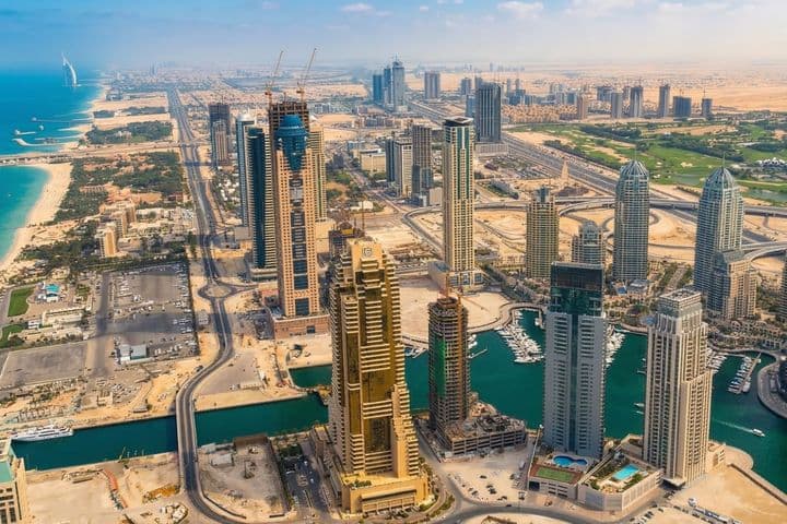Aerial view of early Dubai Marina with skyscrapers under construction, winding roads, and a canal, set against a backdrop of sand and the distant coastline.