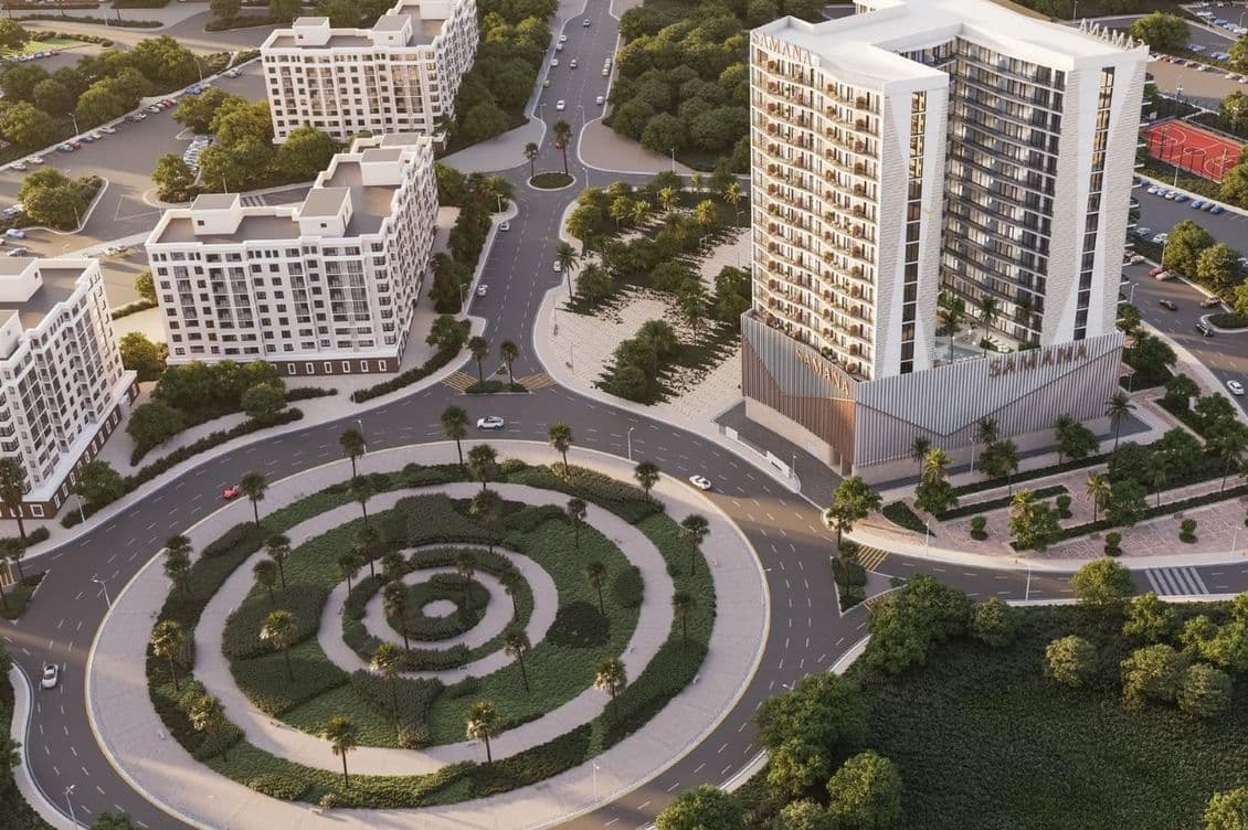Aerial view of a modern urban area with a tall building marked "Samana," surrounded by roads, smaller buildings, and green spaces.