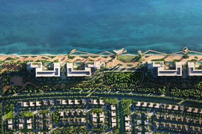 Aerial view of a coastal development with turquoise sea, beachfront promenade, modern white waterfront buildings and tree-lined residential blocks.