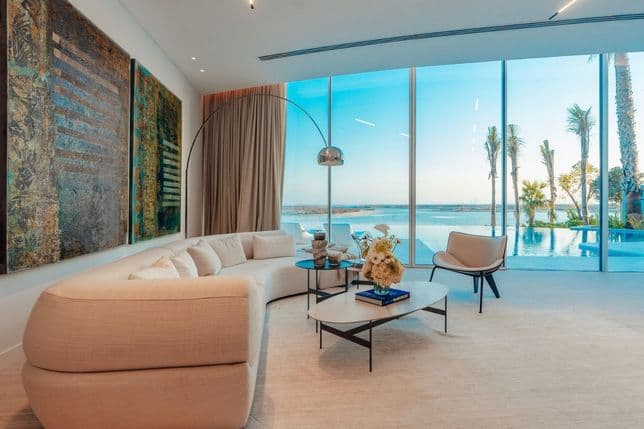 Modern living room with a curved beige sofa, abstract wall art, coffee table, and large windows overlooking a pool and palm trees outside.