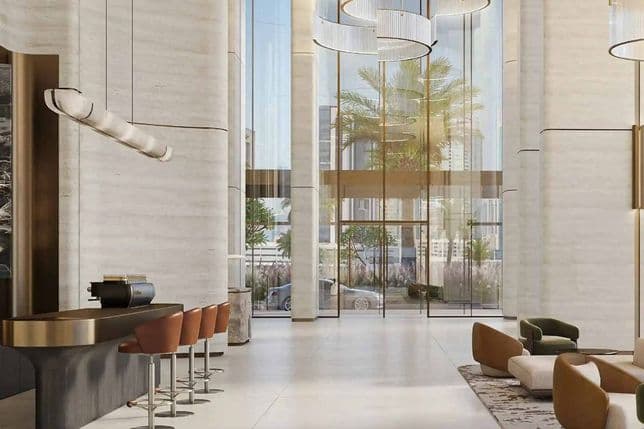 Modern lobby with tall ceilings, stylish light fixtures, a sleek bar with stools, and large windows showing outdoor greenery and palm trees.