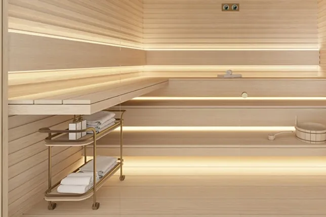 Bright wooden sauna with two-tier benches, warm backlit shelves, a towel cart with towels, and a bucket with a ladle. KI-generiertes Bild