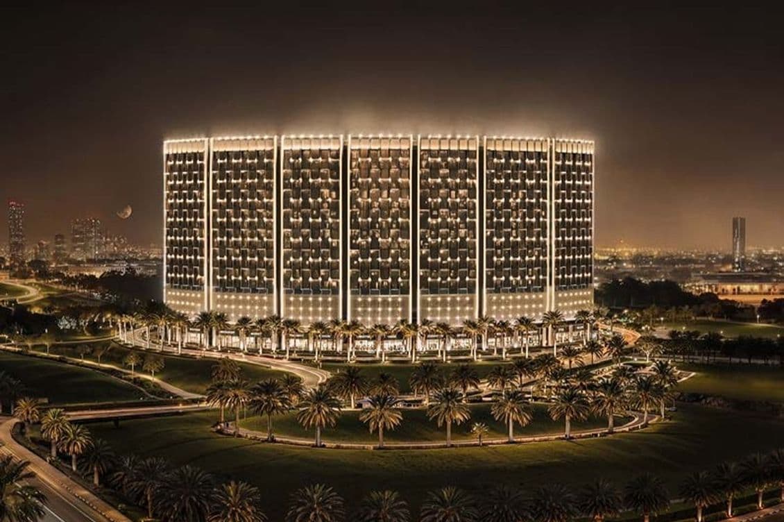 Night view of a large, illuminated building with grid-patterned windows, encircled by palm-lined roads; distant city lights in the background. AI-generated image