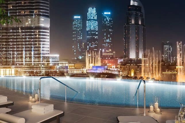 Rooftop infinity pool overlooking a city skyline at night, with illuminated skyscrapers and a lit fountain in the background.