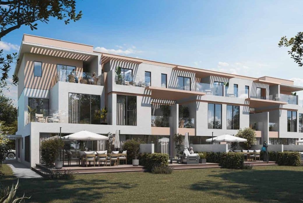 Modern three-story apartment building with balconies, large windows, terrace seating, and a grassy foreground under a clear blue sky.