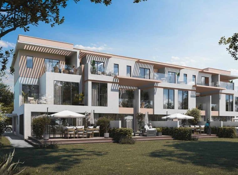 Modern three-story apartment building with balconies, large windows, terrace seating, and a grassy foreground under a clear blue sky.