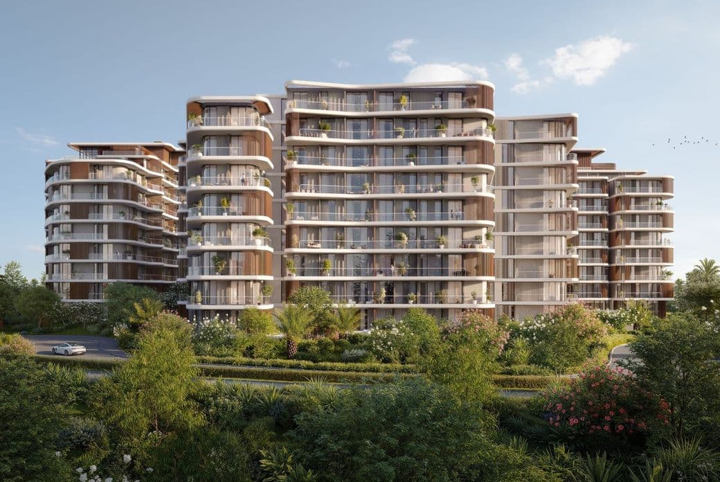 Modern multi-story building with curved balconies, surrounded by lush greenery and a clear blue sky. A car is visible on the road in front.