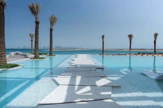 Infinity pool with a white stone walkway, surrounded by palm trees, overlooking a distant city skyline and calm blue sea under a clear sky.