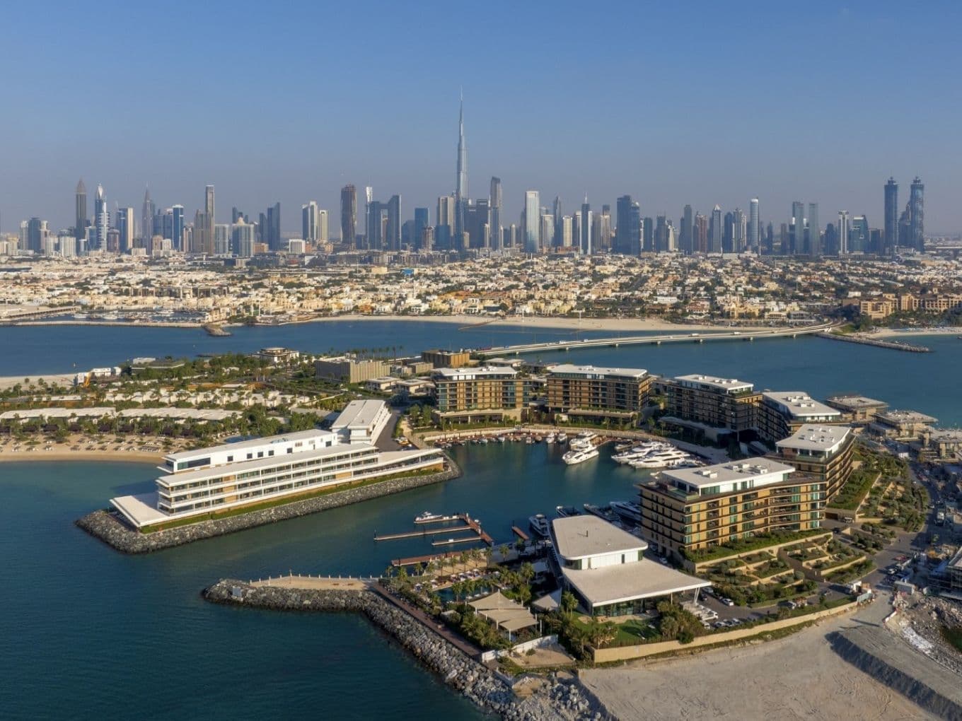 Luftaufnahme der Skyline von Dubai mit dem Burj Khalifa im Hintergrund und Gebäuden am Wasser im Vordergrund unter klarem blauem Himmel.