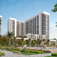 Modern apartment complex with two tall buildings, surrounded by greenery. A playground with swings and slides is in the foreground under a blue sky.
