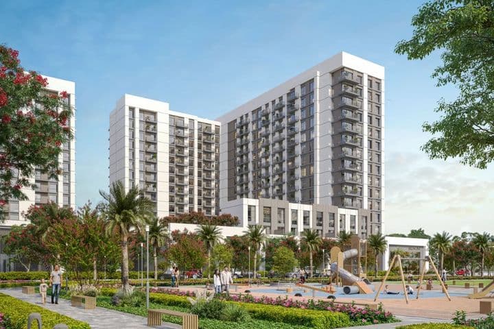 Modern apartment complex with two tall buildings, surrounded by greenery. A playground with swings and slides is in the foreground under a blue sky.
