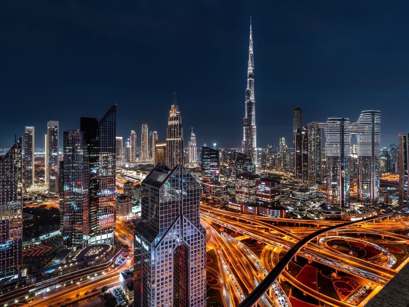 A vibrant nighttime cityscape of Dubai, featuring illuminated skyscrapers, including the Burj Khalifa, and intricate highway patterns.