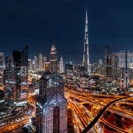A vibrant nighttime cityscape of Dubai, featuring illuminated skyscrapers, including the Burj Khalifa, and intricate highway patterns.