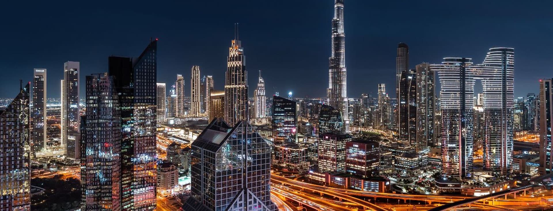 A vibrant nighttime cityscape of Dubai, featuring illuminated skyscrapers, including the Burj Khalifa, and intricate highway patterns.