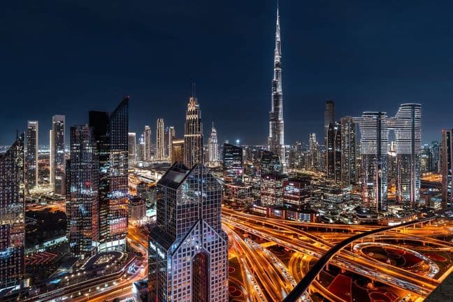 A vibrant nighttime cityscape of Dubai, featuring illuminated skyscrapers, including the Burj Khalifa, and intricate highway patterns.