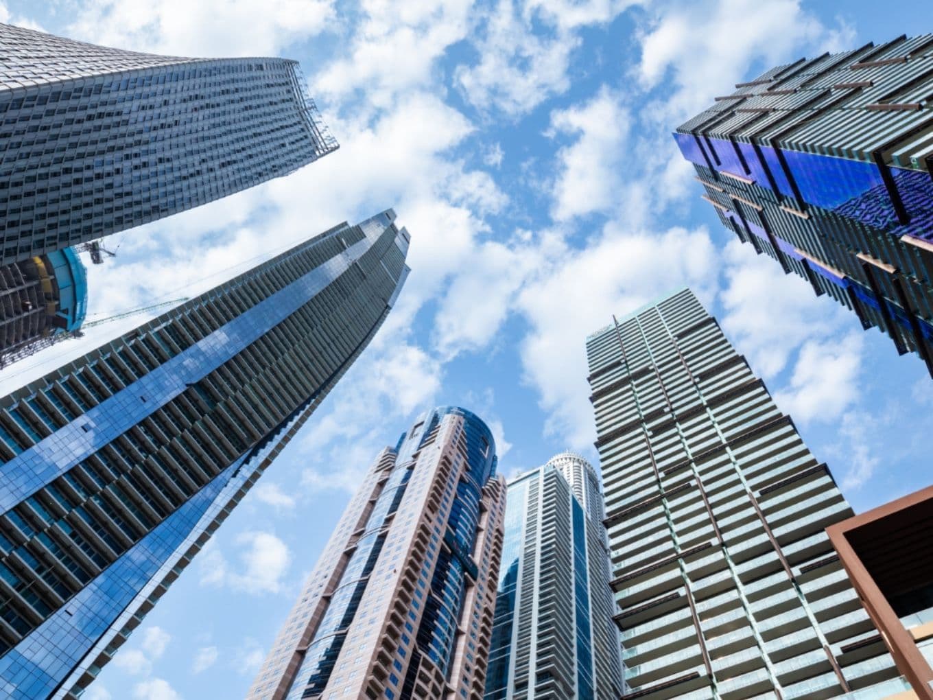 Upward view of modern skyscrapers with various designs against a blue sky dotted with clouds, creating a dramatic urban skyline.