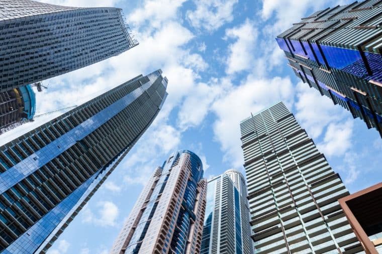 Upward view of modern skyscrapers with various designs against a blue sky dotted with clouds, creating a dramatic urban skyline.