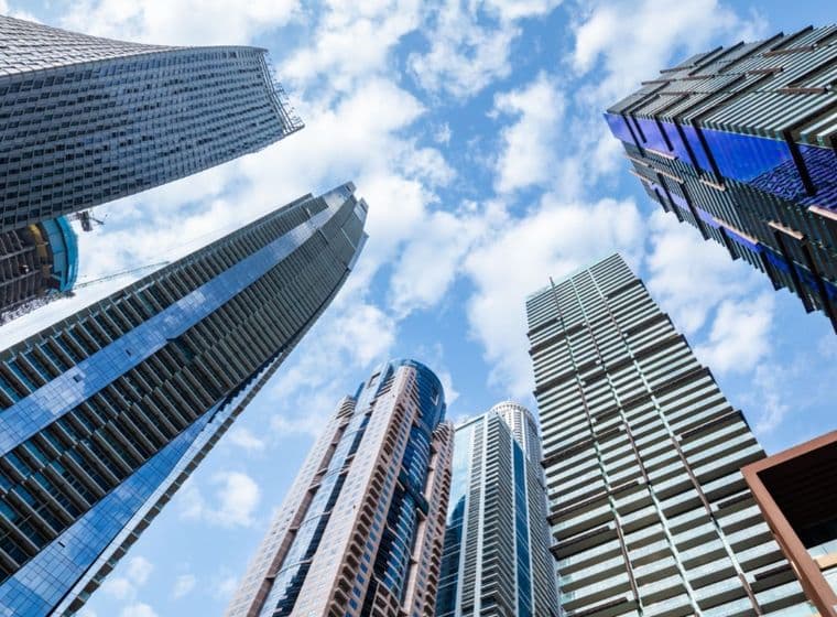 Upward view of modern skyscrapers with various designs against a blue sky dotted with clouds, creating a dramatic urban skyline.