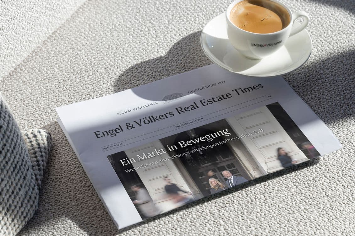 Zeitung mit dem Titel „Engel & Völkers Real Estate Times” auf einem hellen Teppich, neben einer weißen Espressotasse auf einer passenden Untertasse.