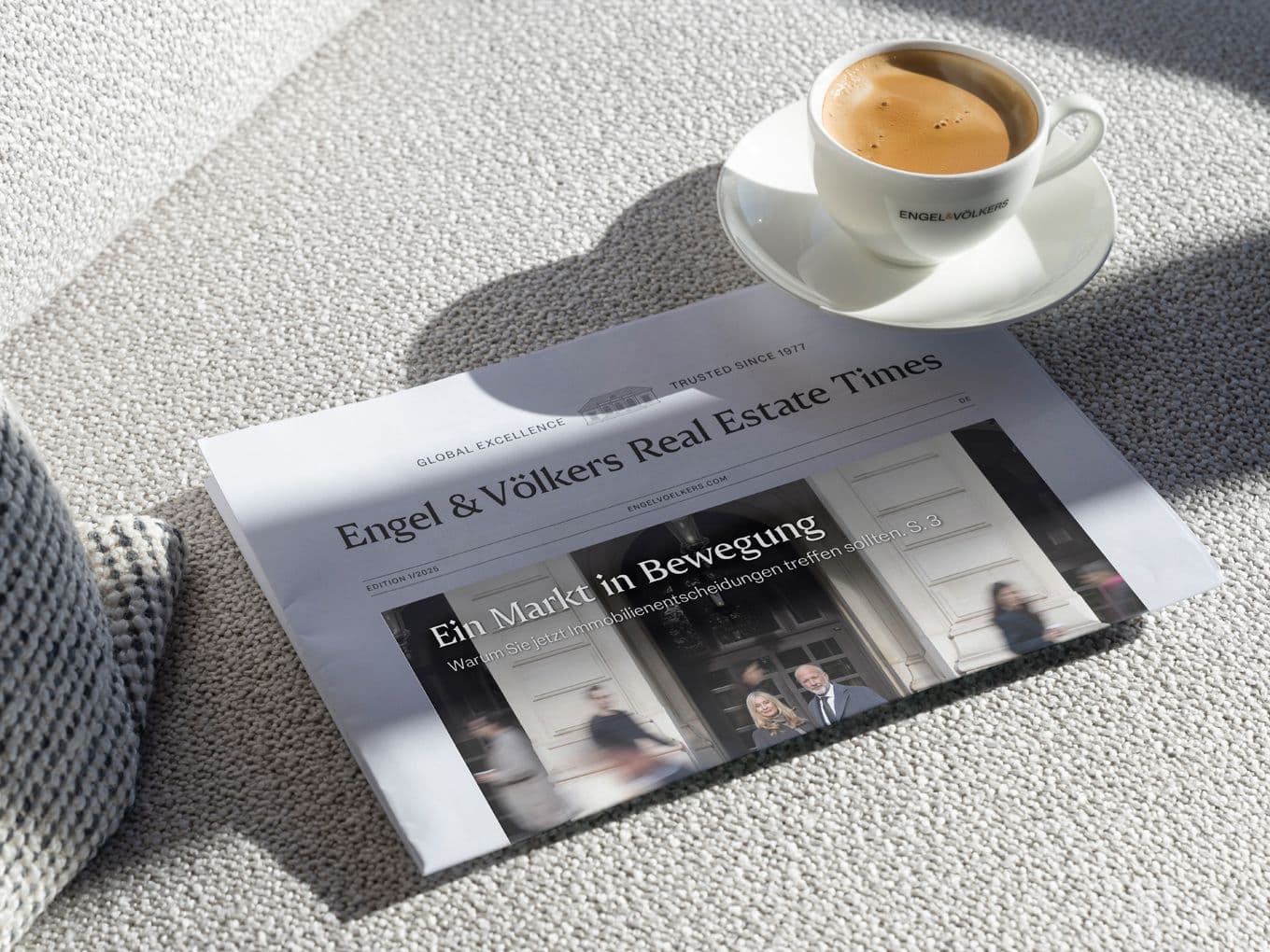 Newspaper titled "Engel & Völkers Real Estate Times" on a light carpet, next to a white cup of espresso placed on a matching saucer.