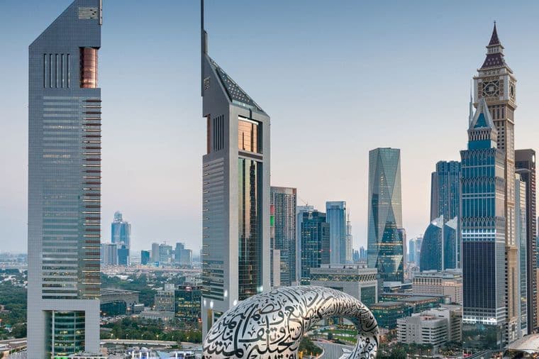 Dubai skyline with futuristic glass skyscrapers; foreground features a large white ring covered in Arabic calligraphy.