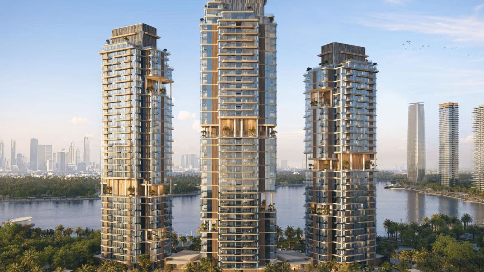 Three modern high-rise buildings with glass facades stand near a waterfront, surrounded by greenery, under a clear blue sky.
