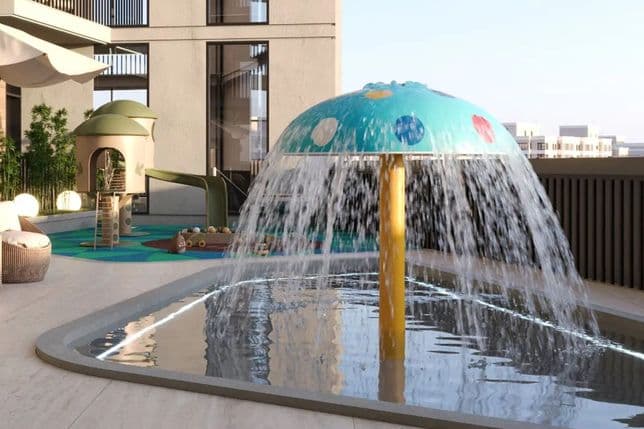 Children's play area with water feature shaped like a mushroom umbrella, slides, and climbing structures, surrounded by city buildings.
