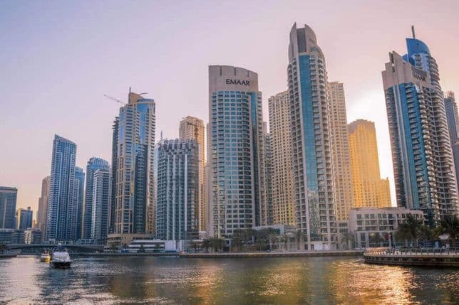 Skyline of modern skyscrapers reflecting in the water at sunset, with a boat on the river in the foreground.