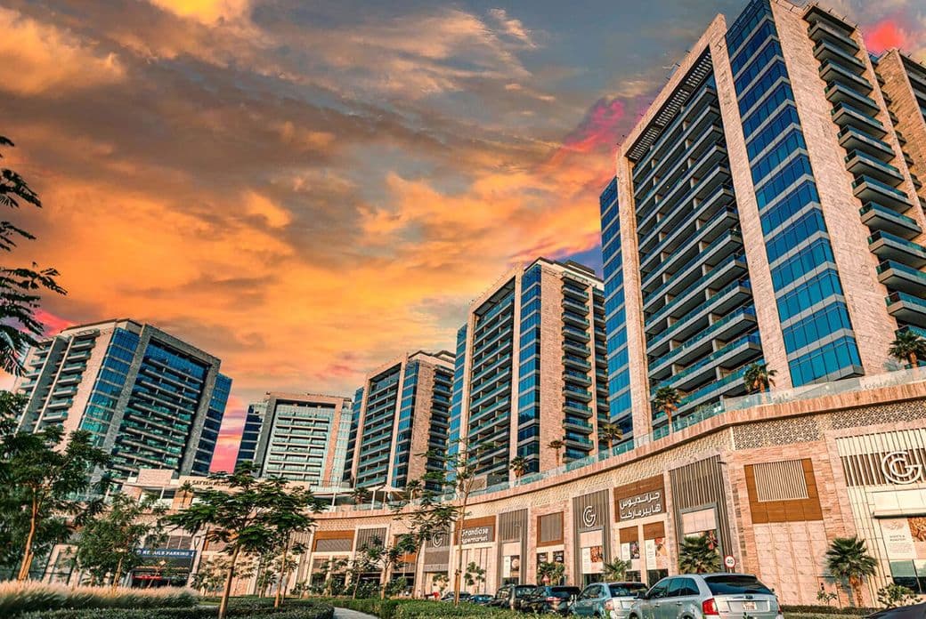Street-level view of Al Sayyah Residence in Arjan, Dubai, during sunset, featuring modern high-rise apartment buildings with glass balconies above a retail podium that includes Grandiose Supermarket and other shops. The sky glows with warm orange and pink hues, while cars and lush landscaping line the road in the foreground
