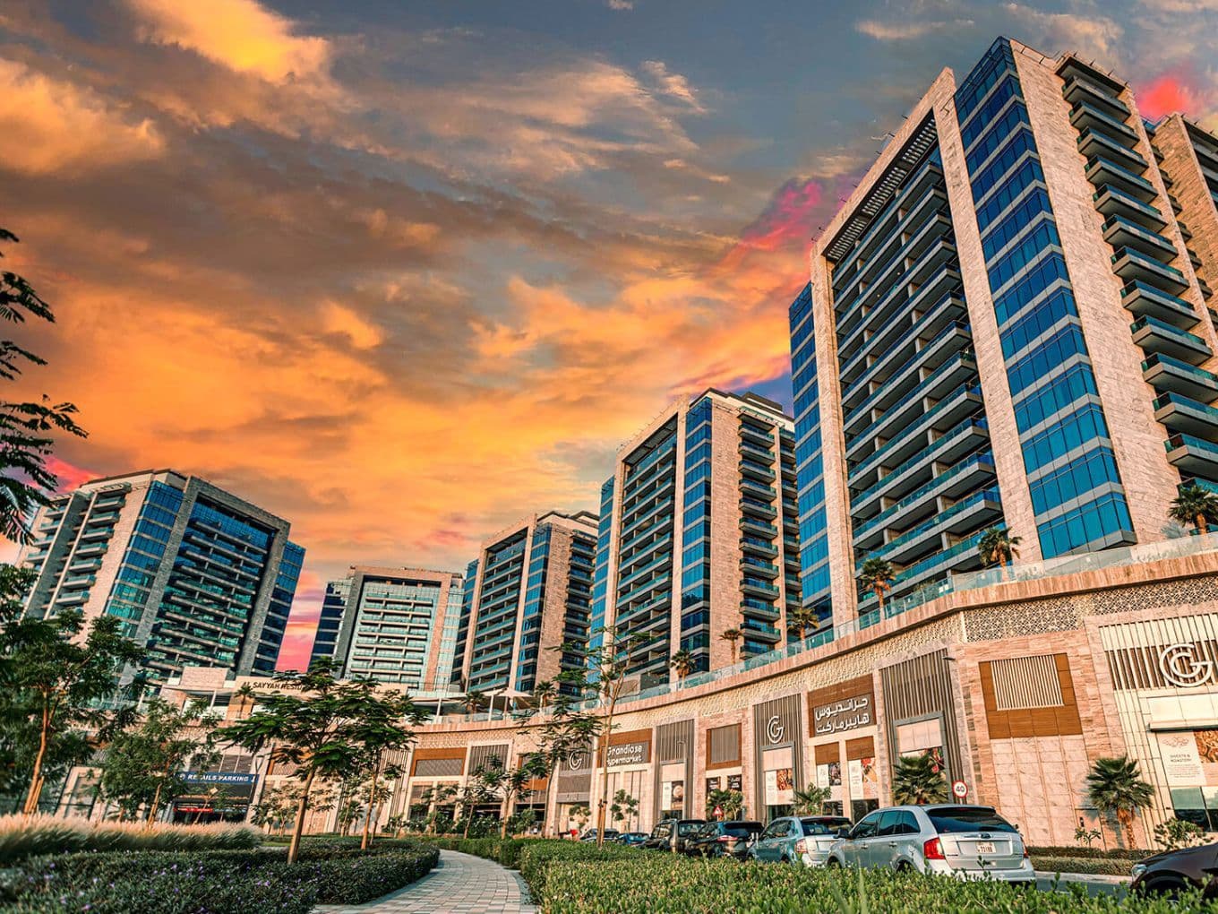 Street-level view of Al Sayyah Residence in Arjan, Dubai, during sunset, featuring modern high-rise apartment buildings with glass balconies above a retail podium that includes Grandiose Supermarket and other shops. The sky glows with warm orange and pink hues, while cars and lush landscaping line the road in the foreground