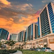 Street-level view of Al Sayyah Residence in Arjan, Dubai, during sunset, featuring modern high-rise apartment buildings with glass balconies above a retail podium that includes Grandiose Supermarket and other shops. The sky glows with warm orange and pink hues, while cars and lush landscaping line the road in the foreground