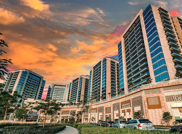 Street-level view of Al Sayyah Residence in Arjan, Dubai, during sunset, featuring modern high-rise apartment buildings with glass balconies above a retail podium that includes Grandiose Supermarket and other shops. The sky glows with warm orange and pink hues, while cars and lush landscaping line the road in the foreground