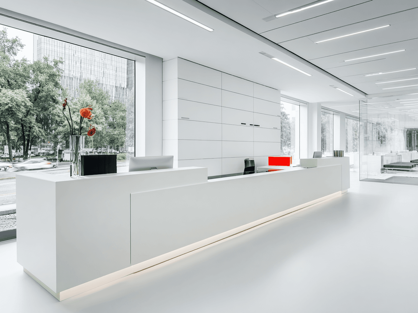 Modern, minimalist office reception with a sleek white desk, flower vase, and large windows offering a view of trees and city buildings.