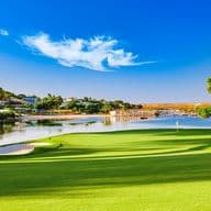 Malerischer Golfplatz mit üppig grünem Fairway, Sandbunker und einem See. Bäume und Häuser im Hintergrund unter einem klaren blauen Himmel.