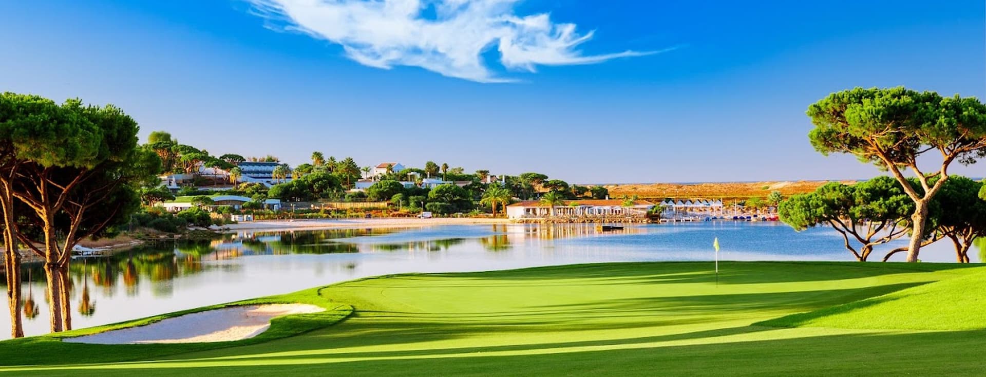 Malerischer Golfplatz mit üppig grünem Fairway, Sandbunker und einem See. Bäume und Häuser im Hintergrund unter einem klaren blauen Himmel.