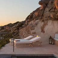 Sonnenuntergangsblick über ein ruhiges Meer von einer felsigen Klippenterrasse mit Lounge-Stühlen, einem kleinen Pool und einem gedeckten Esstisch.