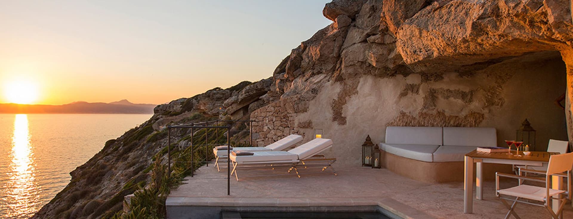 Sonnenuntergangsblick über ein ruhiges Meer von einer felsigen Klippenterrasse mit Lounge-Stühlen, einem kleinen Pool und einem gedeckten Esstisch.