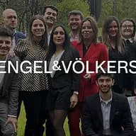 Een groep Engel & Völkers makelaars poseert buiten, lachend naar de camera. De makelaars dragen zakelijke kleding in zwart, rood en grijs.