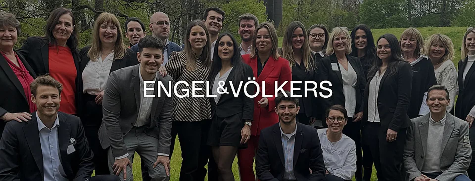 Un groupe d'agents immobiliers Engel & Völkers posent en extérieur, souriant à la caméra. Les agents portent des tenues professionnelles en noir, rouge et gris.