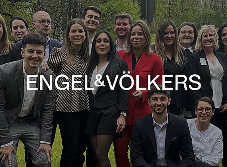 Een groep Engel & Völkers makelaars poseert buiten, lachend naar de camera. De makelaars dragen zakelijke kleding in zwart, rood en grijs.