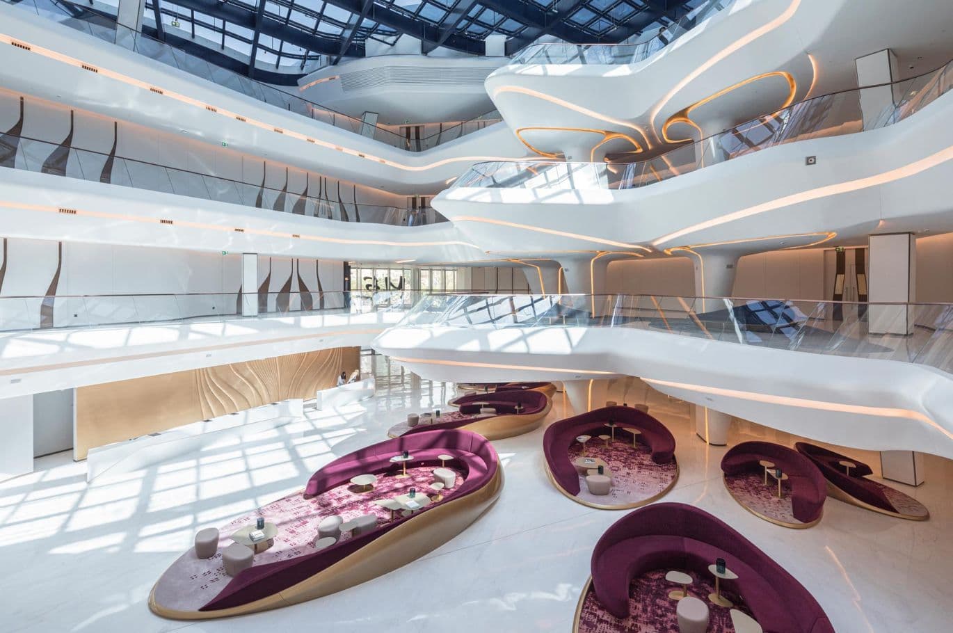 Futuristic atrium interior of The Opus by Omniyat in Business Bay, featuring flowing white architectural lines with integrated lighting, glass balconies, and modern lounge seating areas with curved purple sofas and patterned rugs