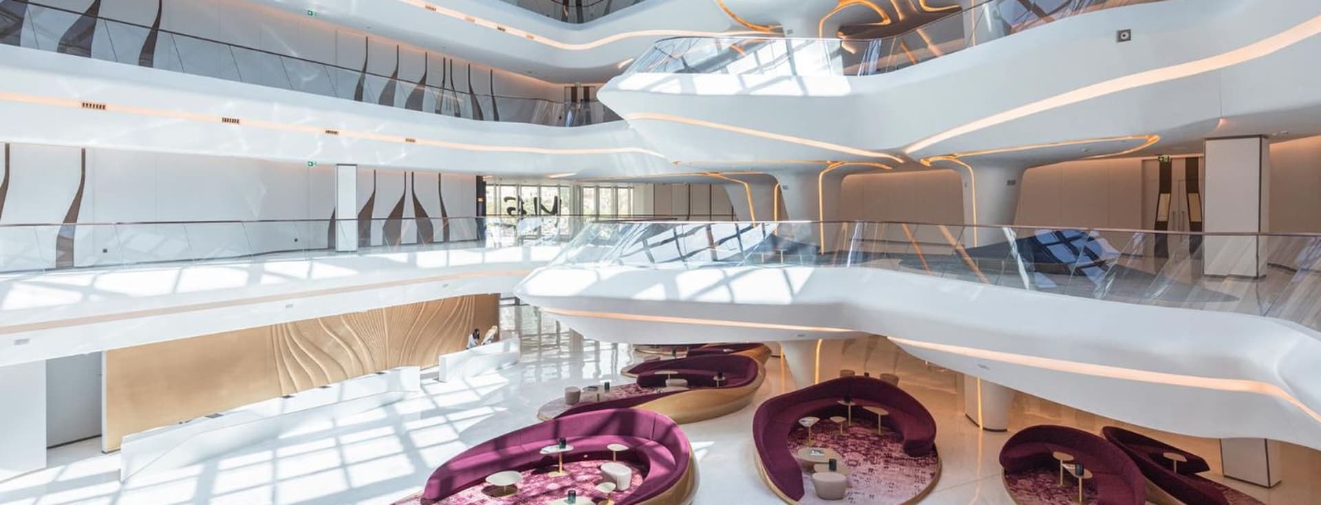 Futuristic atrium interior of The Opus by Omniyat in Business Bay, featuring flowing white architectural lines with integrated lighting, glass balconies, and modern lounge seating areas with curved purple sofas and patterned rugs