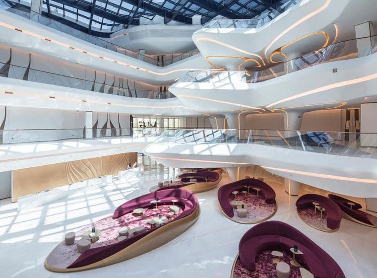 Futuristic atrium interior of The Opus by Omniyat in Business Bay, featuring flowing white architectural lines with integrated lighting, glass balconies, and modern lounge seating areas with curved purple sofas and patterned rugs