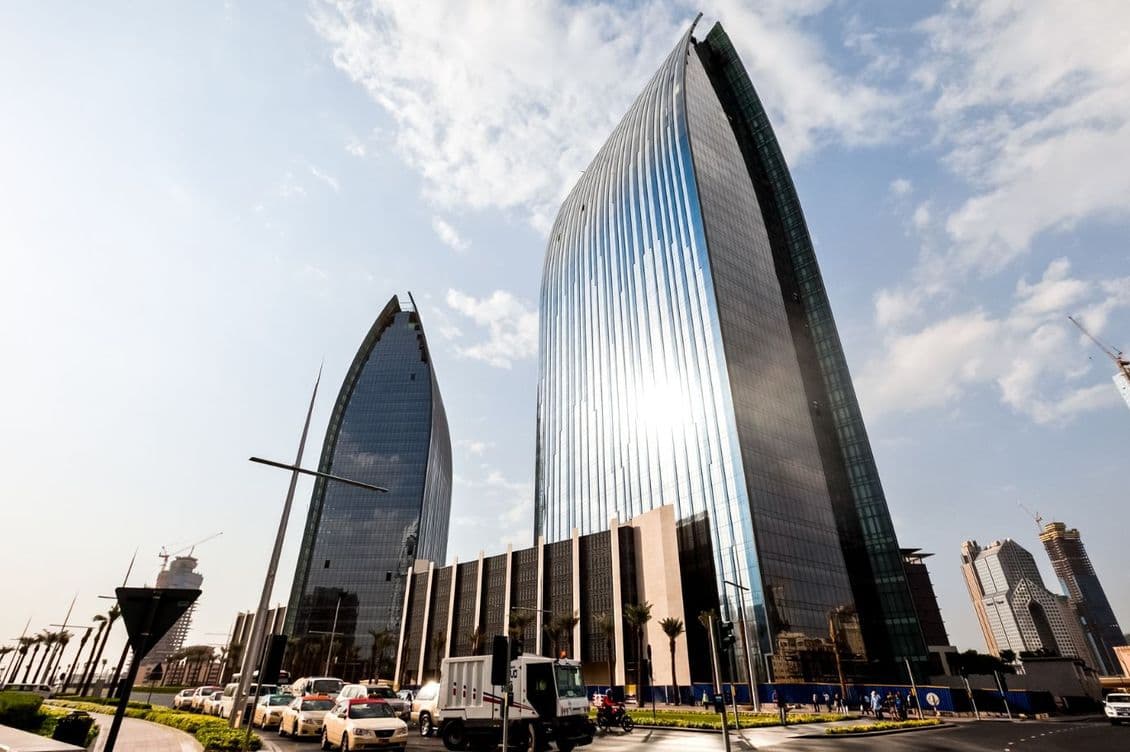 Two modern, reflective skyscrapers with curved designs stand against a partly cloudy sky. Traffic and streetlights are visible in the foreground.