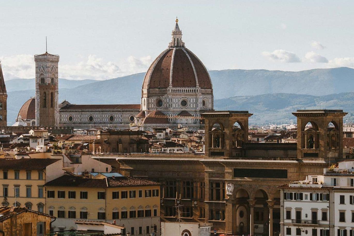 View of Florence with the iconic Florence Cathedral, featuring its large dome, surrounded by historic buildings and distant mountains.
