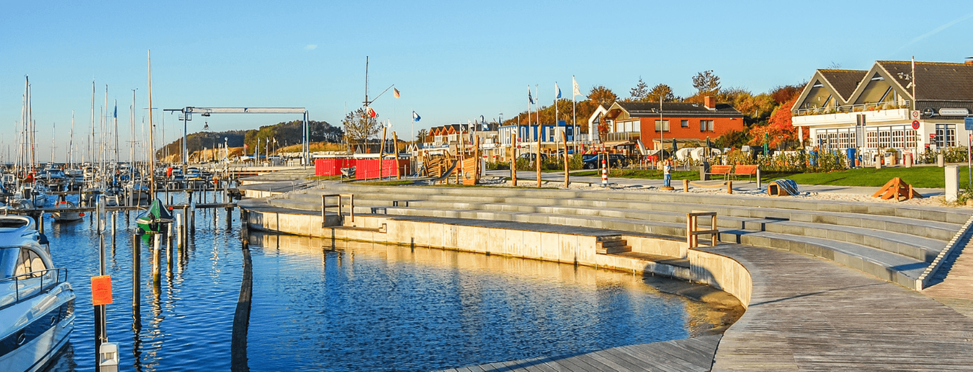 Scenic view of a marina with boats docked, a curved wooden walkway, and colorful buildings under a clear blue sky.