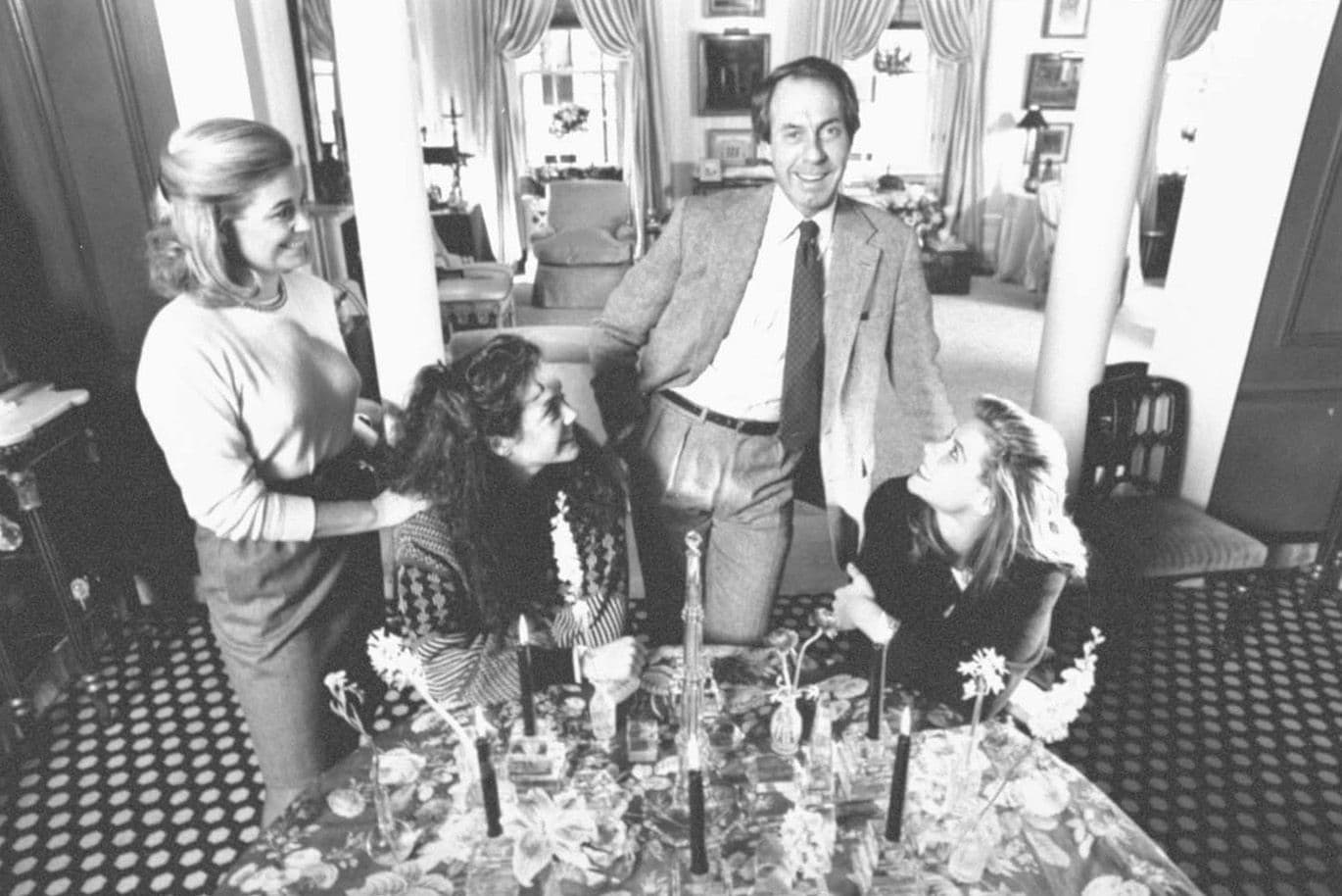 A black-and-white photo of four people gathered around a dining table with flowers and candles in a well-decorated room.