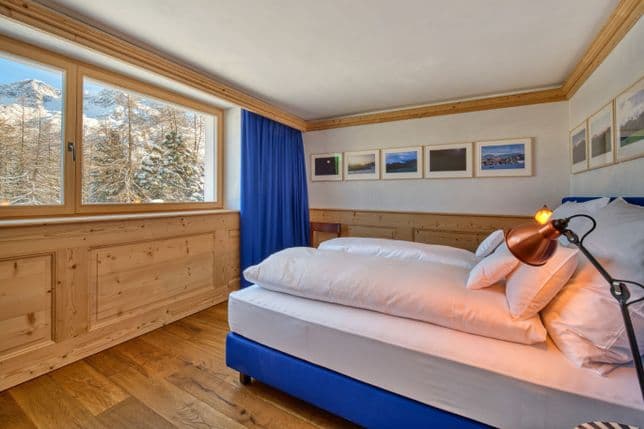 Gemütliches Schlafzimmer mit Holzvertäfelung, blauem Bett und Vorhängen, weißer Bettwäsche und großem Fenster mit Blick auf verschneite Kiefern und Berge.