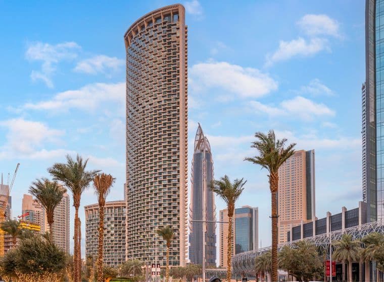 Tall curved Burj Vista tower in Dubai surrounded by palm trees wrapped in decorative lights, modern skyscrapers, and a blue sky with scattered clouds.