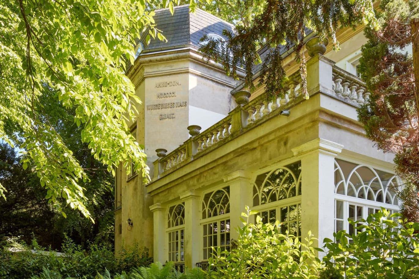 Historic building with arched windows, surrounded by lush greenery and trees, bathed in sunlight.